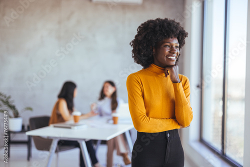 Фотография Beautiful young grinning professional Black woman in office, folded arms and confident expression as other workers hold a meeting in background