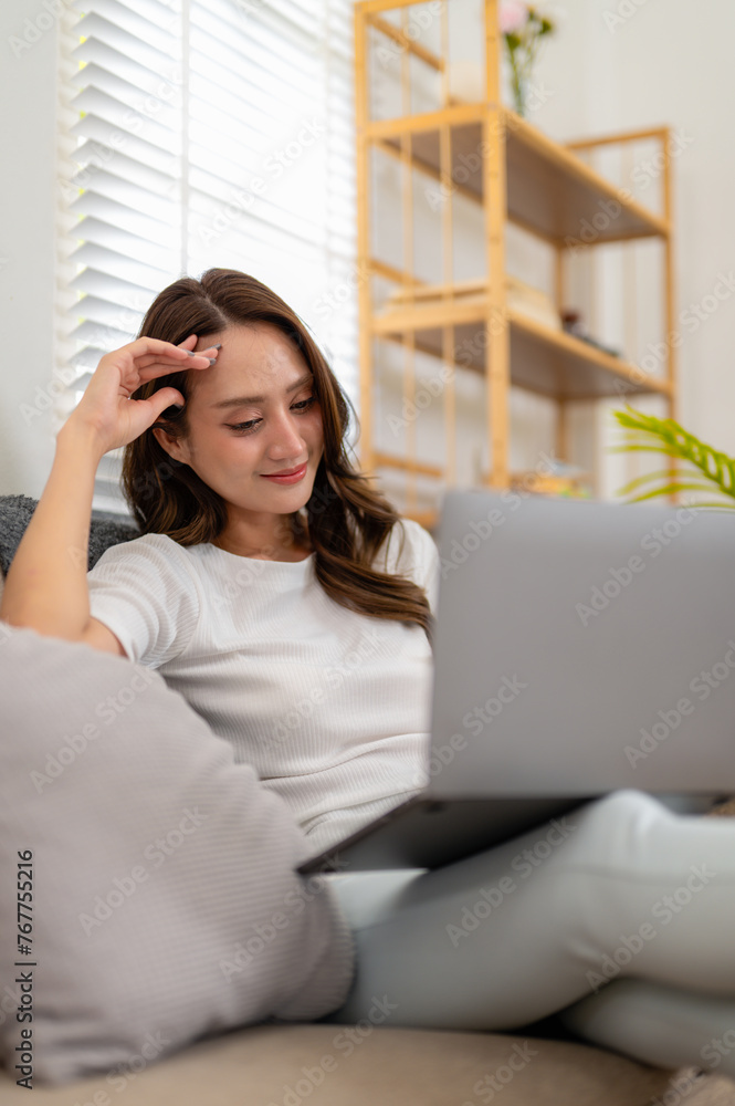 A young woman enjoys a leisurely day, comfortably using her laptop while reclining on a couch at home. A young woman enjoys a leisurely day, comfortably using her laptop while reclining on a couch at home.