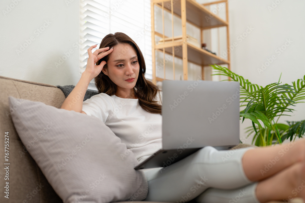 Fototapeta premium A young woman enjoys a leisurely day, comfortably using her laptop while reclining on a couch at home.