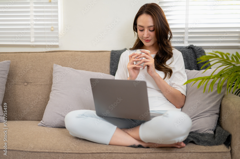 A young woman enjoying a relaxing day. Using a laptop comfortably while reclining and sipping coffee on the sofa at home A young woman enjoying a relaxing day. Using a laptop comfortably while reclining and sipping coffee on the sofa at home
