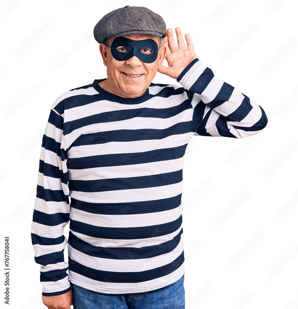 Senior handsome man wearing burglar mask and t-shirt smiling with hand over ear listening an hearing to rumor or gossip. deafness concept. Senior handsome man wearing burglar mask and t-shirt smiling with hand over ear listening an hearing to rumor or gossip. deafness concept.