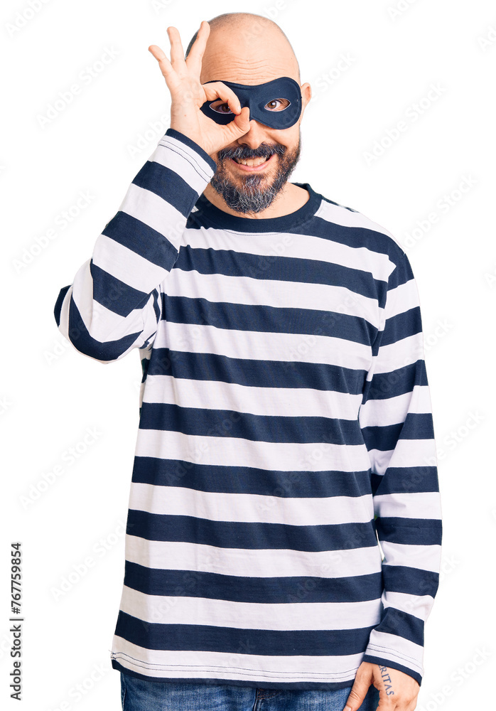 Young handsome man wearing burglar mask doing ok gesture with hand smiling, eye looking through fingers with happy face. Young handsome man wearing burglar mask doing ok gesture with hand smiling, eye looking through fingers with happy face.