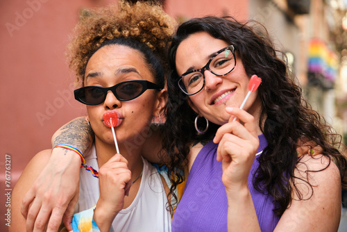 Lesbian couple are holding heart lollipops and smiling at camera celebrating Pride Day.