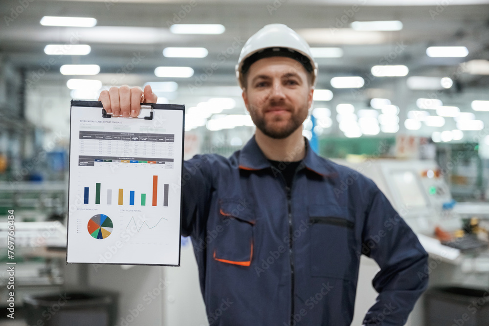 Plan with graphs in hand. Factory worker is indoors with hard hat Plan with graphs in hand. Factory worker is indoors with hard hat