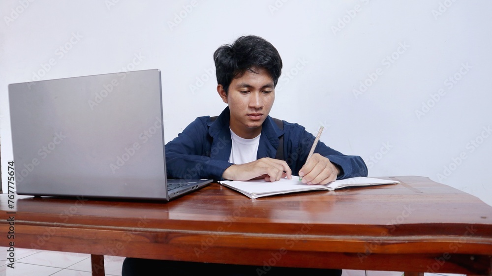 Handsome young Asian male student studying at a table using a book and laptop Handsome young Asian male student studying at a table using a book and laptop