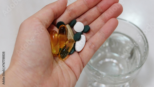 Hand holding assorted supplement pills and capsules. With a glass of water in the background.