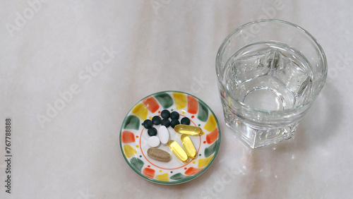 A small plate with various health supplement pills and capsules. On a marble surface, with a glass of water beside with plate.