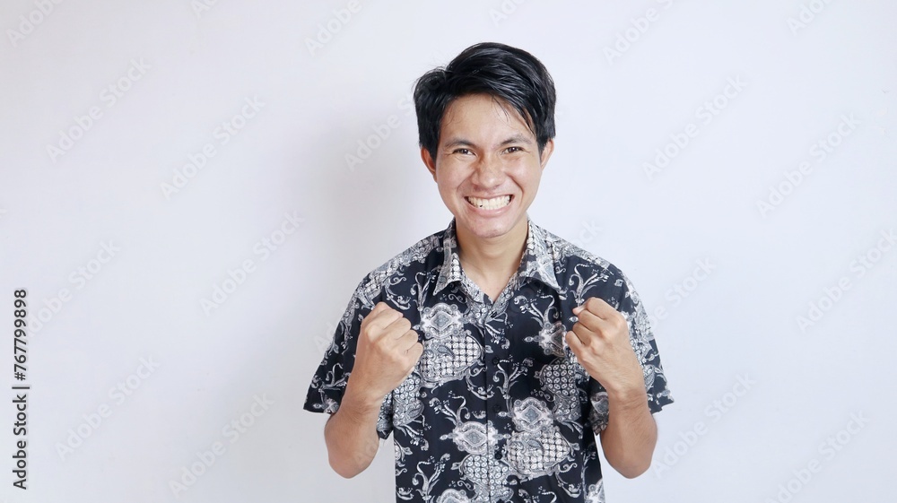 Excited handsome Asian young man wearing batik gesturing celebrating victory and raising Excited handsome Asian young man wearing batik gesturing celebrating victory and raising