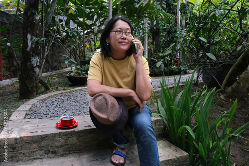 Asian middle-aged woman sitting alone in the garden making a phone call with her smartphone photo