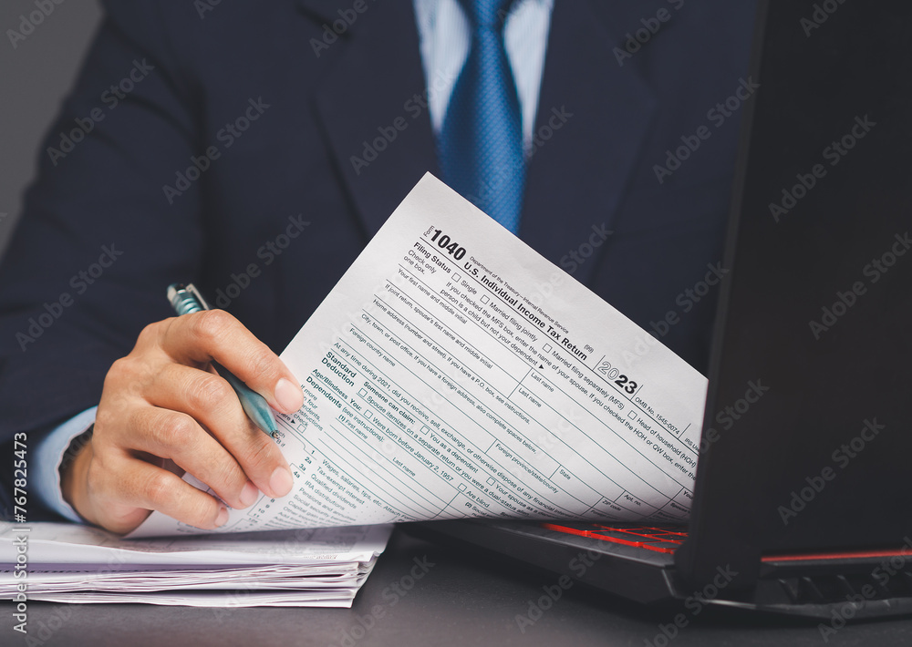 A businessman holding a pen and filling in a tax form with a laptop. A businessman holding a pen and filling in a tax form with a laptop.