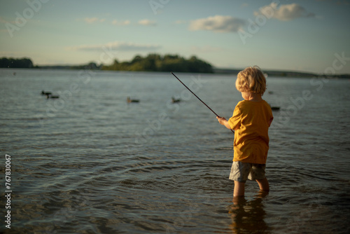 fair-haired little three-year-old boy plays on the lake in the summer, a child plays fisherman on the lake, children's activity on the water, children's imitation of adults, children's curiosity 