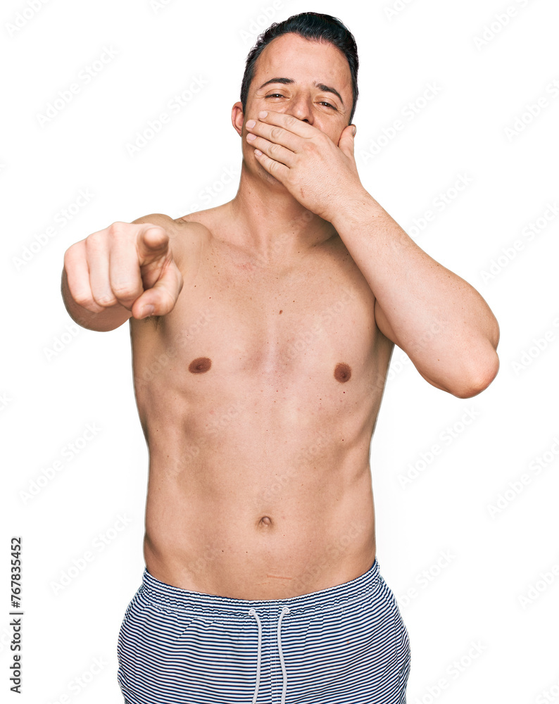 Handsome young man wearing swimwear shirtless laughing at you, pointing finger to the camera with hand over mouth, shame expression