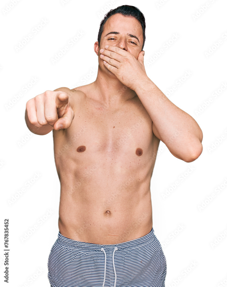 Handsome young man wearing swimwear shirtless laughing at you, pointing finger to the camera with hand over mouth, shame expression Handsome young man wearing swimwear shirtless laughing at you, pointing finger to the camera with hand over mouth, shame expression