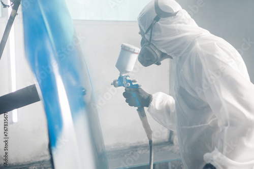 Male Industrial worker sprays paint using airbrush gun on part of car wears in protective mask and overall clothes in workshop garage.
