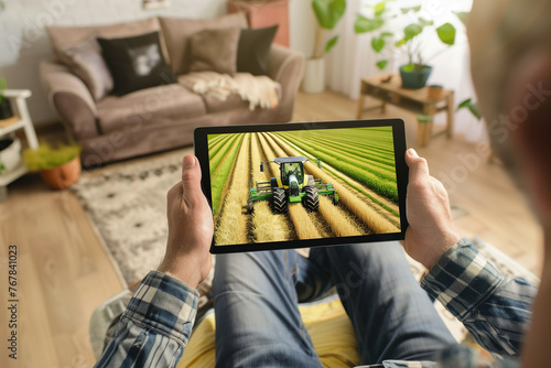 Close up of farmer hands holding digital tablet device and watching tractor in wheat field. Farmer is sitting in living room. Modern technology and harvesting concept