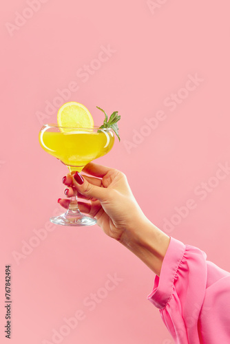 female hand holding vibrant citrus, tropical cocktail decorated with lemon slice and mint leaves against pastel pink background. Concept of alcohol, drink, party, degustation, holiday. Copy space for photo