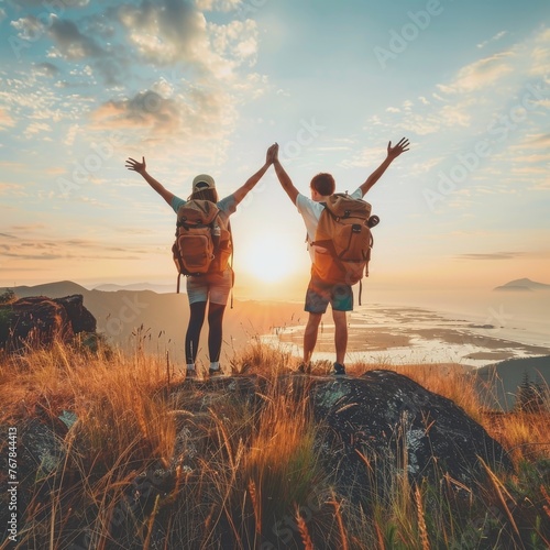 A young man and a young woman stand on top of a hill with arms outstretched at sunset.