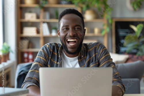 Cheerful young man with stubble looking at laptop screen joyfully raising his hands. Handsome hipster completed successful project, made a lucrative deal. Freelance, creative work and life concept. photo