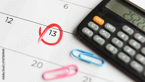 A calendar, paper clips and a calculator are on the table. The date on the calendar – the 13th – is circled with a red marker. Macro.