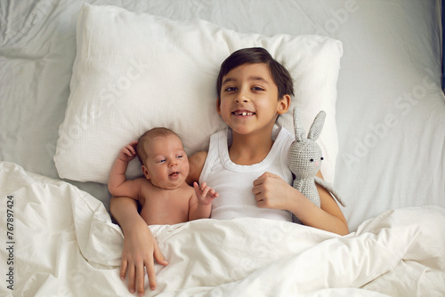cheerful portrait of a boy of seven years old lying with his sister baby on a pillow on a white big bed with a crocheted toy hare , light from the window.
