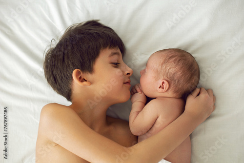 boy seven years old lying with his sister baby in a nappy on his stomach on a white big bed, light from the window