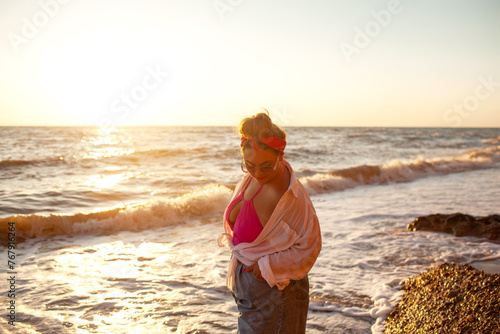 A beautiful plus size woman dressed solid  pink swimsuit, jeans shorts, sunglasses and white shirt walks along the seashore against the sand beach and sea or ocean during the sunset. 