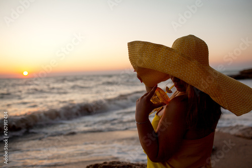 A beautiful plus size woman dressed straw yellow maxi hat and sundress walks along the seashore against the sand beach and sea or ocean during the sunset