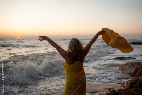 A beautiful plus size woman dressed straw yellow maxi hat and sundress walks along the seashore against the sand beach and sea or ocean during the sunset