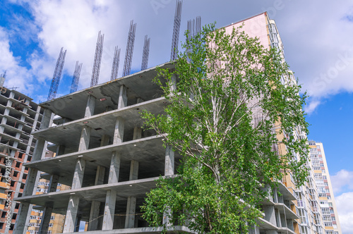 The frame structure of a high-rise building made of reinforced concrete structures. A tree on the background of the frame of a building under construction. Reinforcement with metal bars