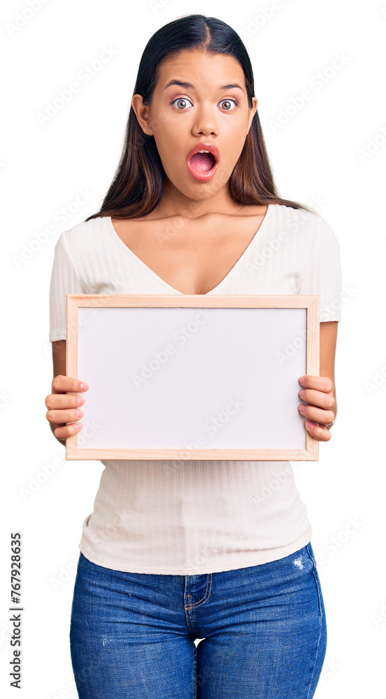 Young beautiful latin girl holding empty white chalkboard scared and amazed with open mouth for surprise, disbelief face Young beautiful latin girl holding empty white chalkboard scared and amazed with open mouth for surprise, disbelief face