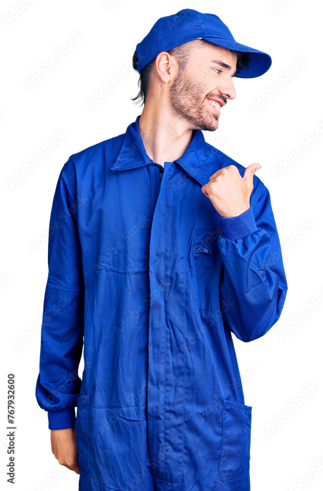 Young hispanic man wearing painter uniform smiling with happy face looking and pointing to the side with thumb up. Young hispanic man wearing painter uniform smiling with happy face looking and pointing to the side with thumb up.
