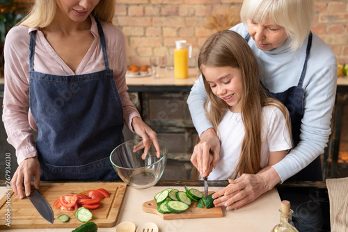 Front shot of family that prepare vegetables for salad. Grandmother showing how to cut cucumber into slices to her granddaughter in cozy kitchen. Three generations spend time together while cooking