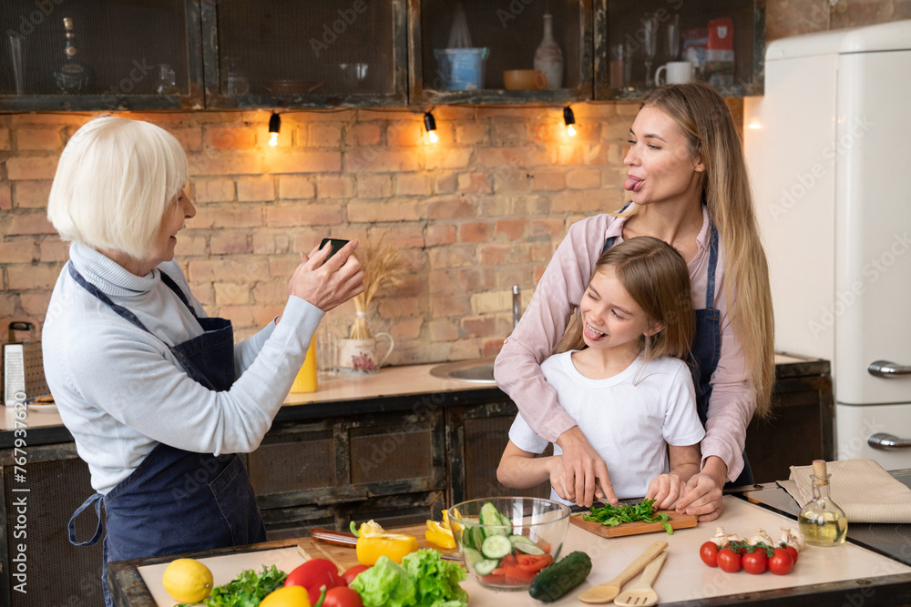 Happy senior woman taking selfie with her daughter and granddaughter in kitchen. Cheerful female and little girl posing while grandmother taking self portrait with mobile phone