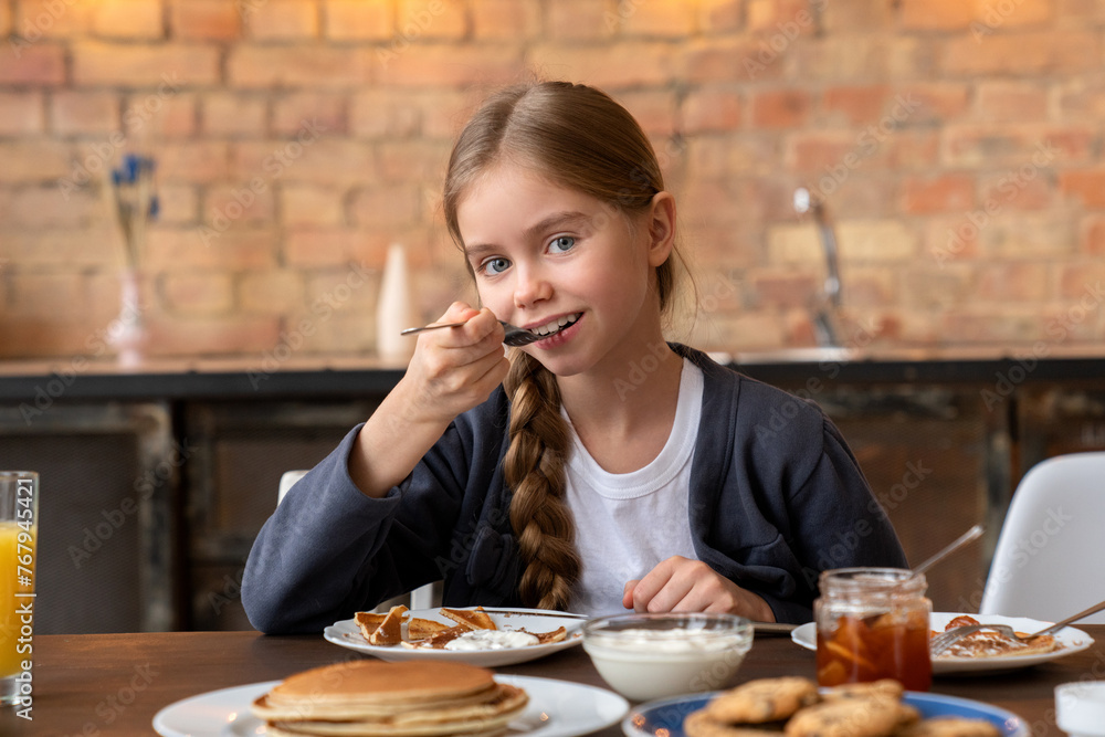 Portrait of happy cute little girl eating pancakes with the spoon on breakfast in kitchen with light bulb on the background. Beautiful girl with blue eyes light hair looking at camera while eating