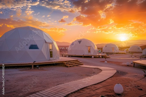 Sunset view of a desert glamping site with futuristic dome tents and wooden walkways.