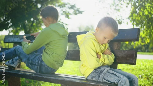 Young boy in bright yellow hoodie sitting on park bench, sulking with arms crossed while his brother absorbed in using smartphone. Children engage with world and each other