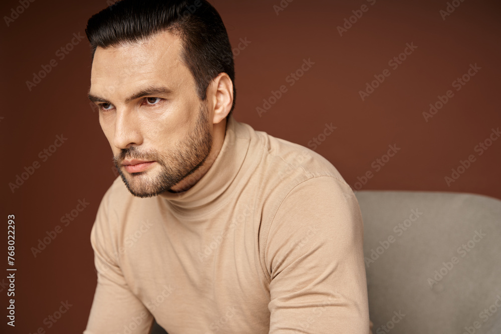 portrait of handsome and serious man in turtleneck sitting on armchair on beige background portrait of handsome and serious man in turtleneck sitting on armchair on beige background