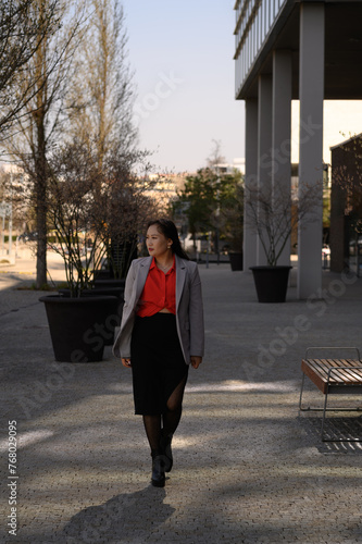 Confident woman in business attire walking through an urban park with modern architecture, wearing a grey blazer, red blouse, black skirt, and boots.