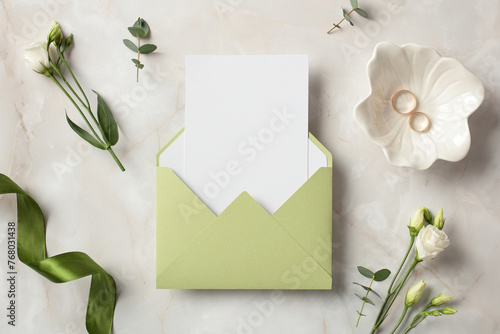 Olive envelope with blank paper card inside, gold rings, green ribbon and flowers on stone background. Wedding invitation card template. Flat lay, top view