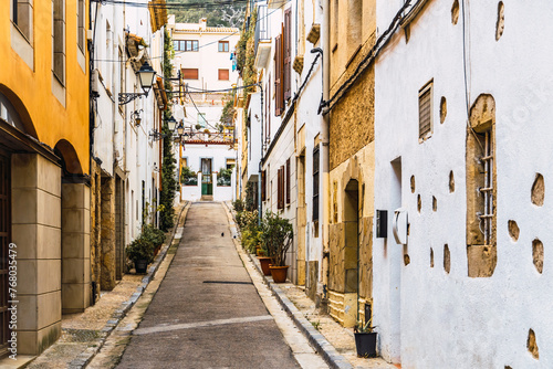 A narrow street with traditional white houses adorned with potted plants, in the old town of the picturesque coastal town of Tossa de Mar, Costa Brava, Spain. © XAVI