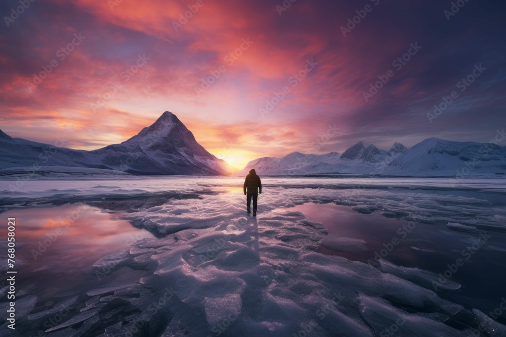 Person standing on a frozen lake with a view of a snow-covered mountain range and a colorful sunset in the background.