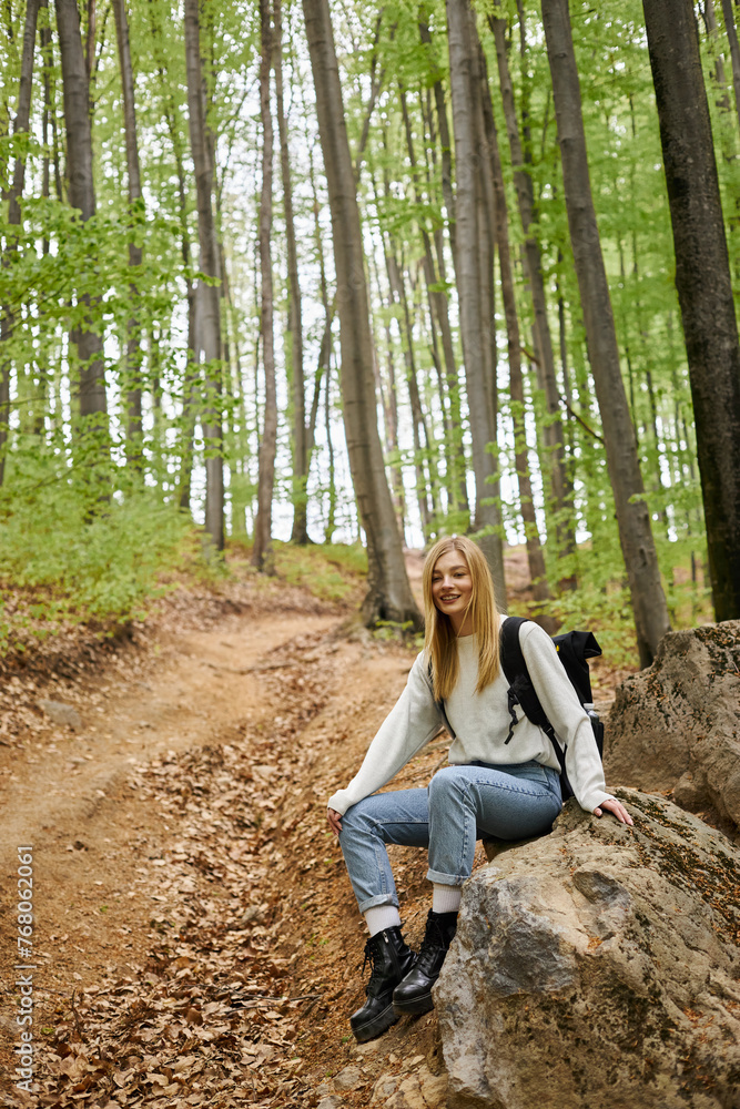 Smiling blonde female hiker wearing backpack, resting on rocks, enjoying the forest view Smiling blonde female hiker wearing backpack, resting on rocks, enjoying the forest view