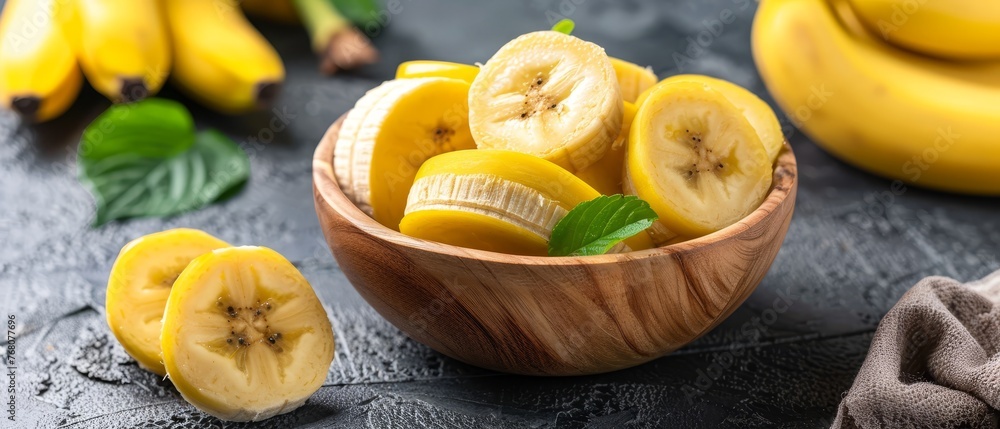 Wooden bowl brimming with sliced bananas near green foliage and bunches of bananas on table Wooden bowl brimming with sliced bananas near green foliage and bunches of bananas on table