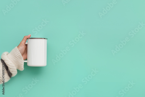 main d'une femme, habillée d'un pull en laine blanc à rayure grise, tenant un mug en métal émaillé en blanc avec un liseré argent, boisson sur fond vert avec espace négatif copy space