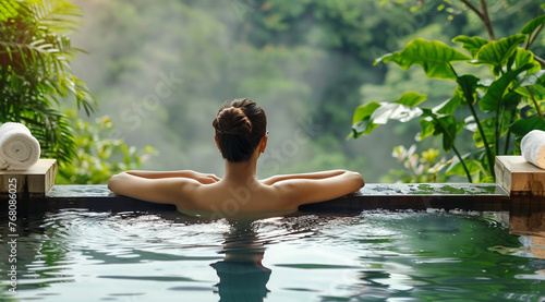 Beautiful young female tourist relaxing in onsen hot spring pool with her back turned,surrounded by the lush greenery. Health and wellness tourism concept.