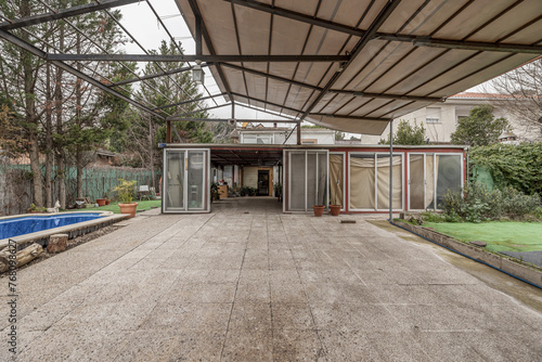 A covered terrace on the plot of a single-family home with a canvas roof on a metal structure and a room with aluminum doors and a vehicle parked inside.