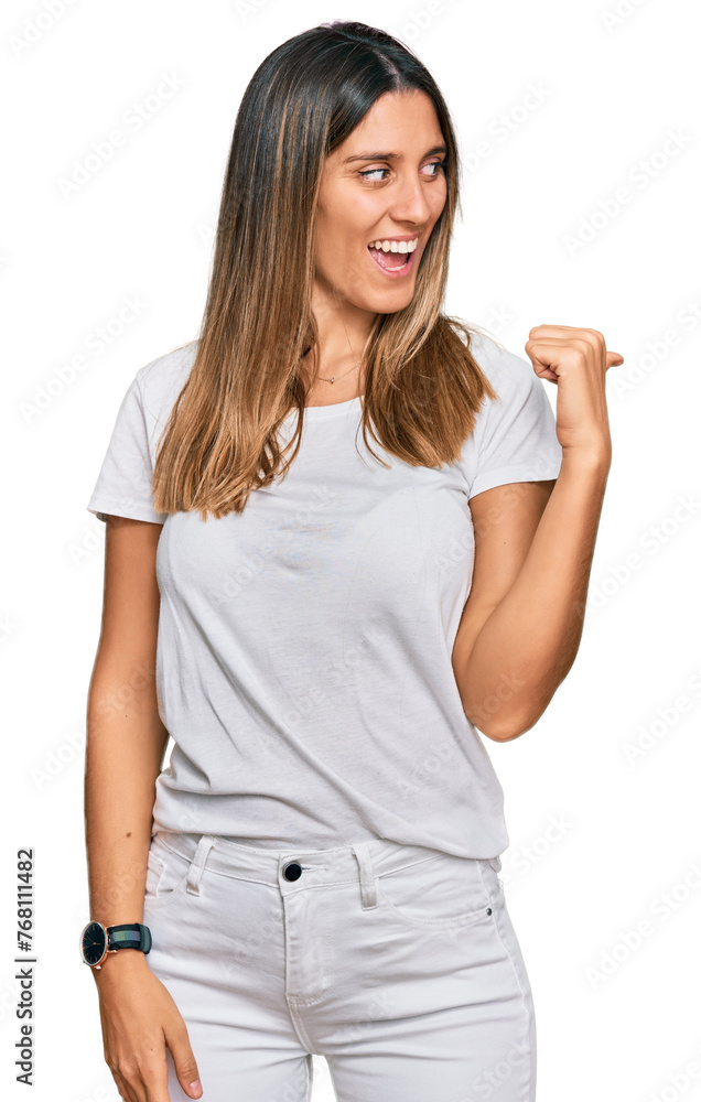 Young woman wearing casual white t shirt smiling with happy face looking and pointing to the side with thumb up. Young woman wearing casual white t shirt smiling with happy face looking and pointing to the side with thumb up.