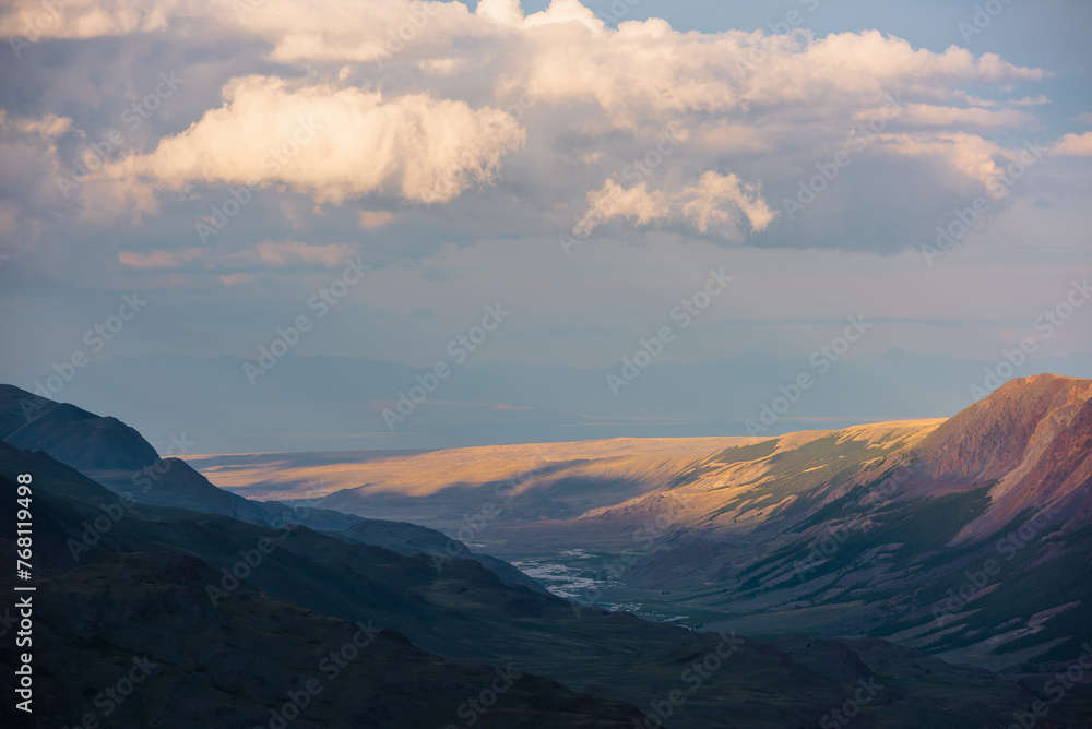 Dramatic aerial top view to golden steppe illuminated by setting sun and mountain range silhouette under clouds in gold sunset tones. Evening alpine landscape under cloudy sky in vivid sunset color.