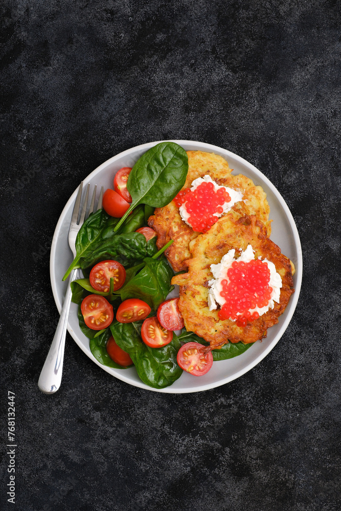 Fried potatoes pancakes with salmon caviar and cream cheese with spinach and tomatoes on black background. Fried potatoes pancakes with salmon caviar and cream cheese with spinach and tomatoes on black background.