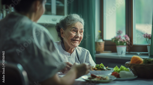  In a hospital ward, a retired woman is visited by a caring nurse, sharing a light moment during lunchtime as they enjoy a meal together